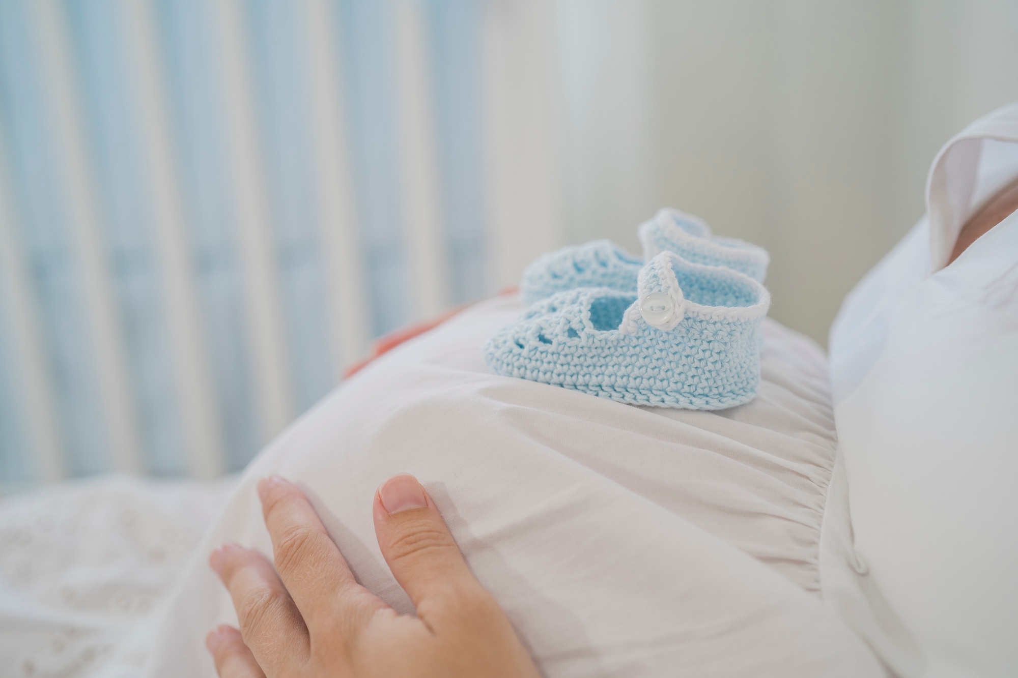 Crop faceless pregnant lady with baby shoes on tummy lying on bed
